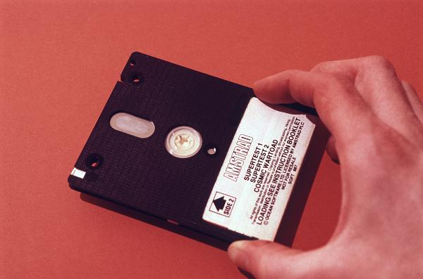 Man holding a 3 inch computer disk in his hand with a specification label over a red background, this type of disk was commonly used with amstrad computers in the 1980's
