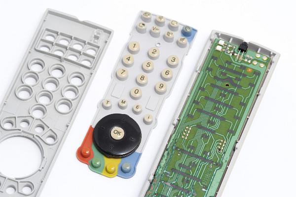 Disassembled remote control lying in three parts showing the case, buttons or keypad and the electronic circuitry. The buttons are formed of flexible rubber and a conductive carbon pad which connects to pads on the circuitboard when the button is pressed